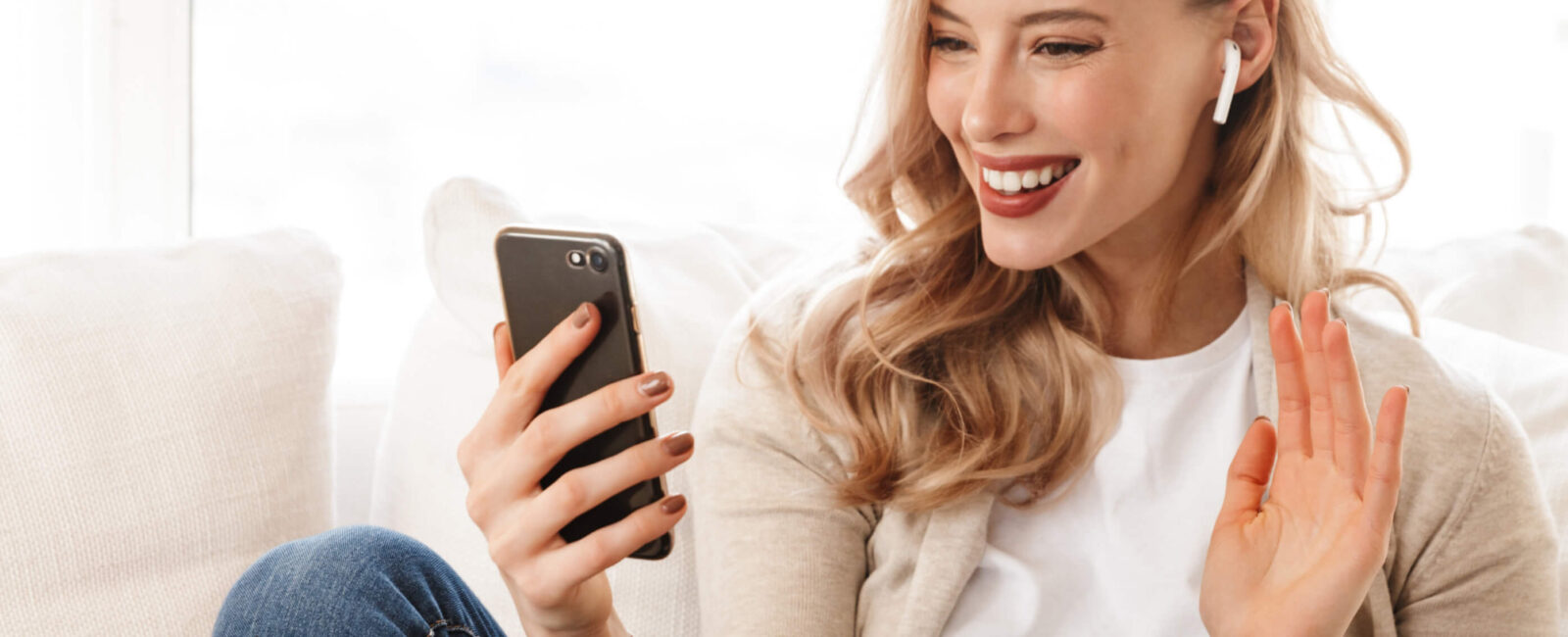 Auf diesem Bild sieht man eine junge Frau, die fröhlich in einen Videocall vertieft ist. Sie sitzt entspannt auf einem Sofa, trägt kabellose Kopfhörer und hält ein Smartphone in der Hand, während sie lächelnd und freundlich in die Kamera winkt. Die Umgebung wirkt hell und einladend. Die Perspektive zeigt ihr Gesicht im Detail, wobei das Licht und die Kameraposition ihre Gesichtszüge schmeichelhaft betonen – ein Aspekt, der oft entscheidend ist, wenn es darum geht, das sogenannte „Videocall Doppelkinn“ zu vermeiden oder zu minimieren. Für diejenigen, die eine dauerhafte Lösung suchen, könnte eine Behandlung wie CoolSculpting, die Fettwegspritze oder Radiofrequenz-Microneedling in Wien in Betracht gezogen werden, um ein Videocall Doppelkinn entfernen.