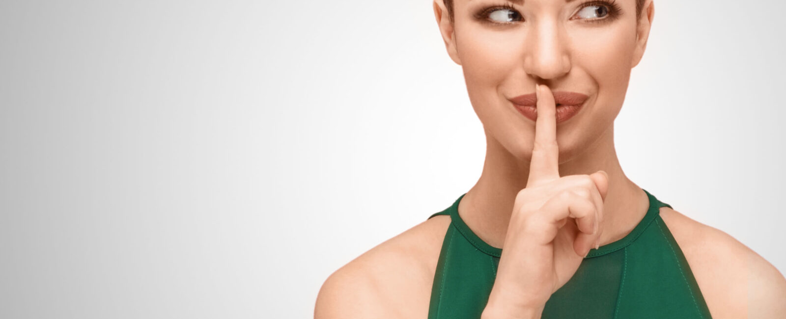 Eine Frau in einem grünen Kleid hält den Finger an ihre Lippen, ein Symbol für Geheimhaltung. Das Bild thematisiert die Verjüngung der Hände und wie Hände das wahre Alter verraten können. Altersflecken und andere Pigmentstörungen lassen sich mit CO2-Laserbehandlungen und Hyaluronsäure-Fillern, wie sie bei Dermacare Wien angeboten werden, effektiv behandeln.