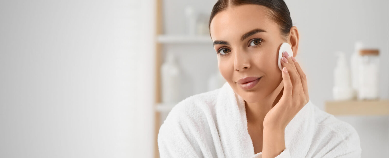 Eine Frau in einem weißen Bademantel pflegt ihre Haut mit einem Wattepad. Sie hat einen frischen, natürlichen Teint und trägt kein Make-up. Im Hintergrund sind Pflegeprodukte zu sehen. Die Bildkomposition vermittelt das Thema Microneedling Pflege und zeigt, dass die Haut nach der Behandlung sanft gereinigt und gepflegt werden sollte. Wichtige Aspekte wie Make-up nach dem Microneedling und eine geeignete Pflegeroutine stehen im Fokus. Dermacare Wien ist ein erfahrener Ansprechpartner für Microneedling und die richtige Nachsorge.