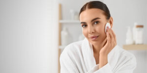 Eine Frau in einem weißen Bademantel pflegt ihre Haut mit einem Wattepad. Sie hat einen frischen, natürlichen Teint und trägt kein Make-up. Im Hintergrund sind Pflegeprodukte zu sehen. Die Bildkomposition vermittelt das Thema Microneedling Pflege und zeigt, dass die Haut nach der Behandlung sanft gereinigt und gepflegt werden sollte. Wichtige Aspekte wie Make-up nach dem Microneedling und eine geeignete Pflegeroutine stehen im Fokus. Dermacare Wien ist ein erfahrener Ansprechpartner für Microneedling und die richtige Nachsorge.