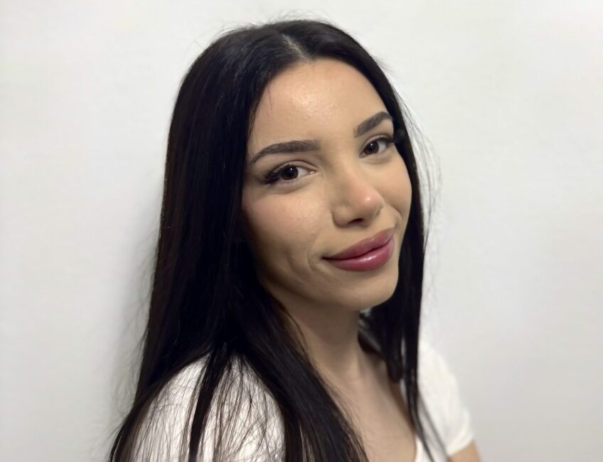 Auf dem Bild ist eine schwarzhaarige junge Frau mit weißem T-shirt zu sehen. Sie arbeitet als Empfangsmitarbeiterin bei Dermacare Wien.