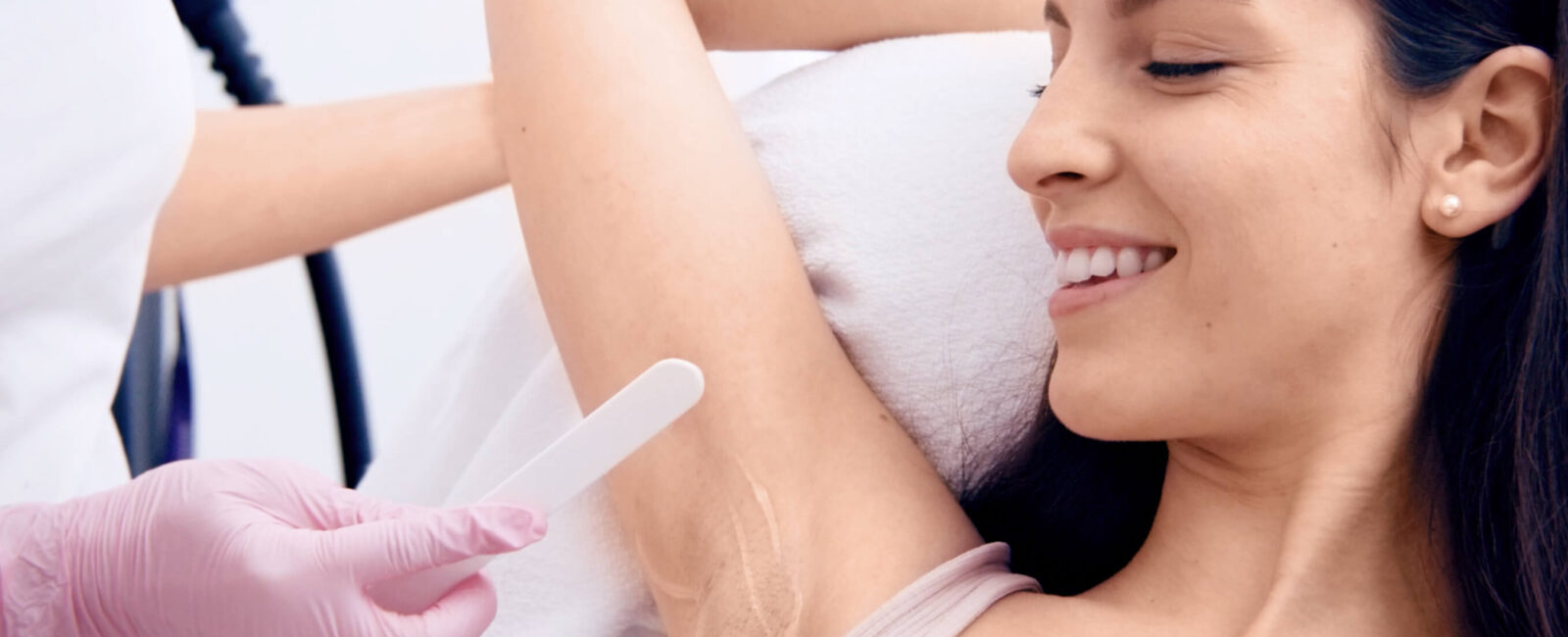Auf dem Bild ist eine junge Frau zu sehen, die entspannt auf einer Behandlungsliege liegt und zufrieden lächelt, während eine Fachkraft mit rosa Handschuhen eine Methode zur Haarentfernung an ihrer Achsel vorbereitet. Dabei handelt es sich um die Vorbereitung auf eine Laser-Haarentfernung eine moderne und effektive Methode zur Haarentfernung, die dauerhaft glatte Haut verspricht. Diese professionelle Behandlung findet bei Dermacare Wien statt, einem spezialisierten Zentrum für ästhetische Medizin. Die glatte Haut der Frau unterstreicht das angestrebte Ergebnis: haarfrei zu sein und sich rundum wohlzufühlen. Das Bild vermittelt ein Gefühl von Hygiene, Vertrauen und moderner Körperpflege.