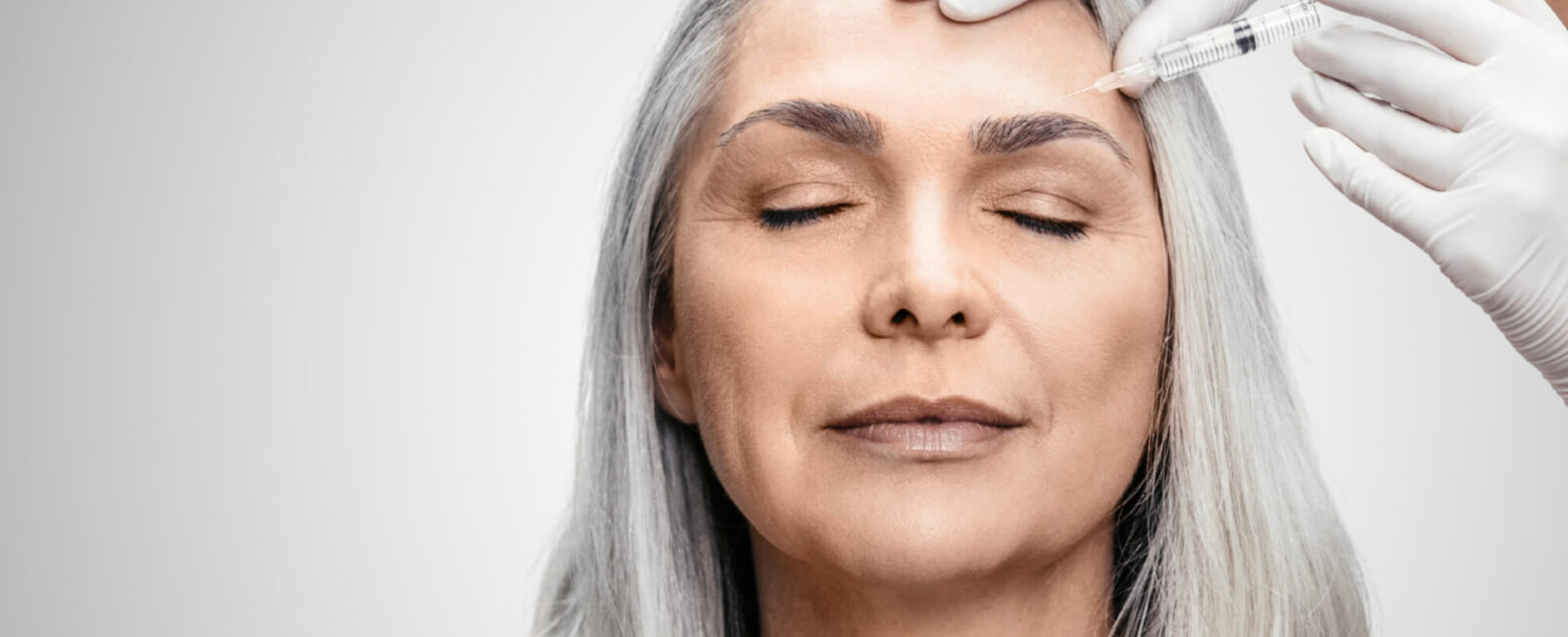 Auf dem Bild sieht man eine Frau mit grauen Haaren, jedoch sehr glatter Haut, besonders zwischen ihren augenbrauen. Sie bekommt in die Stirn gerade eine Faltenunterspritzung von Dermacare Wien. Dermacare beratet und informiert die Patienten immer sorgfältig vor jeder Behandlung auch über die Botox Kosten Zornesfalte.