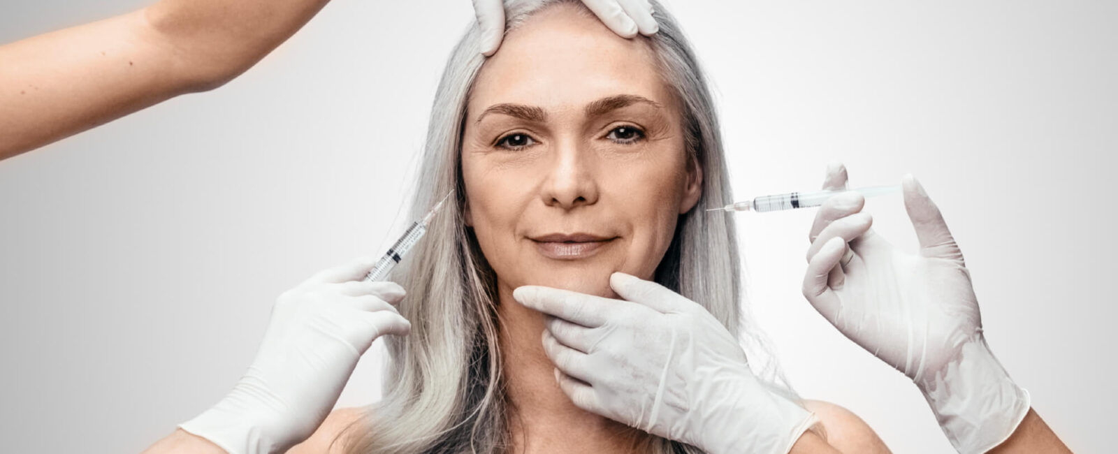 Das Bild zeigt eine elegante, reifere Frau mit silbergrauem Haar, die selbstbewusst und gelassen in die Kamera blickt. Mehrere Hände in weißen medizinischen Handschuhen führen gleichzeitig kosmetische Eingriffe durch – zwei davon setzen Injektionen im Gesichtsbereich an. Diese Szene symbolisiert die professionelle und zugleich sanfte Faltenbehandlung bei Dermacare. Der Fokus liegt auf der Faltenbehandlung mit Hyaluronsäure, die auf natürliche Weise Volumen zurückgibt, Falten glättet und die jugendliche Ausstrahlung der Haut unterstützt. Das neutrale, helle Hintergrunddesign unterstreicht die klinische Präzision und Ästhetik der Behandlung bei Dermacare Wien.