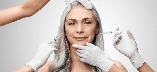 Kombi Faltenbehandlung bei Dermacare – PRP oder Hyaluronsäure? Das Bild zeigt eine elegante, reifere Frau mit silbergrauem Haar, die selbstbewusst und gelassen in die Kamera blickt. Mehrere Hände in weißen medizinischen Handschuhen führen gleichzeitig kosmetische Eingriffe durch – zwei davon setzen Injektionen im Gesichtsbereich an. Diese Szene symbolisiert die professionelle und zugleich sanfte Faltenbehandlung bei Dermacare. Der Fokus liegt auf der Faltenbehandlung mit Hyaluronsäure, die auf natürliche Weise Volumen zurückgibt, Falten glättet und die jugendliche Ausstrahlung der Haut unterstützt. Das neutrale, helle Hintergrunddesign unterstreicht die klinische Präzision und Ästhetik der Behandlung bei Dermacare Wien.