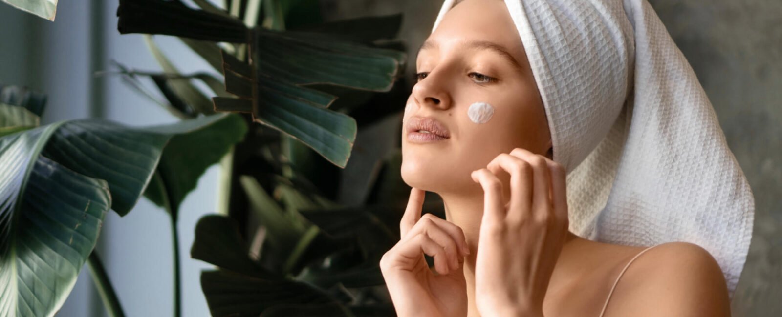 Eine junge Frau mit Handtuchturban trägt sanft eine Pflegecreme auf ihr Gesicht auf. Die Szene vermittelt Frische und Entspannung – perfekt als Symbol für intensive Gesichtspflege und nachhaltige Tiefenreinigung im Sommer. Das Bild passt ideal zum Thema Aquafacial bei Dermacare Wien, das die Haut porentief reinigt und mit Feuchtigkeit versorgt.