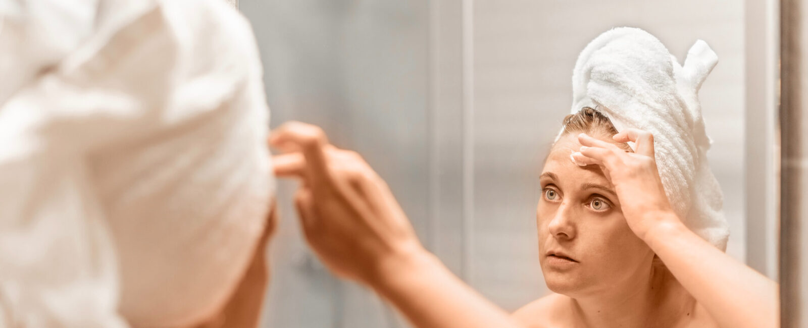 Frau mit Handtuchturban betrachtet sich im Spiegel und berührt ihre Stirn, zum Stirnfalten glätten – Symbol für Anti-Aging- und Hautpflegebehandlungen.