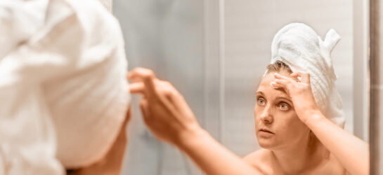 Frau mit Handtuchturban betrachtet sich im Spiegel und berührt ihre Stirn, zum Stirnfalten glätten – Symbol für Anti-Aging- und Hautpflegebehandlungen.