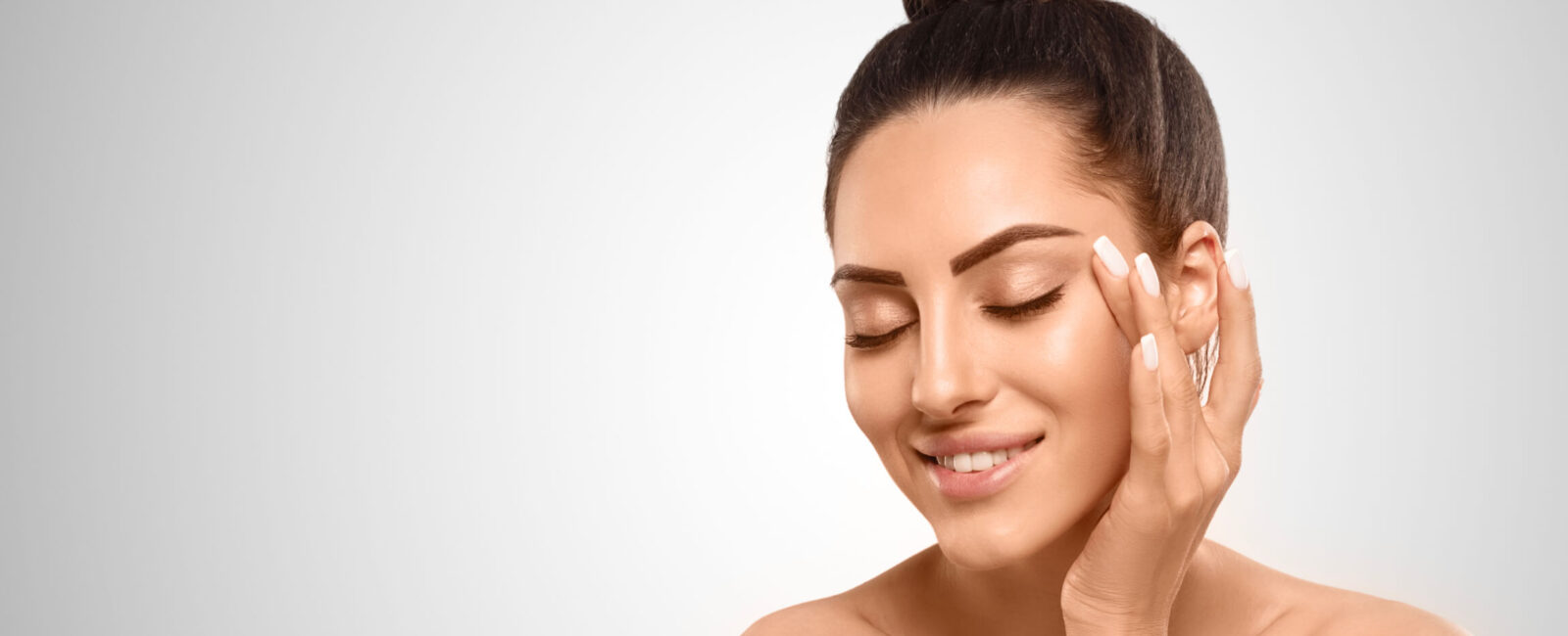 Junge Frau mit gepflegter Haut lächelt entspannt und berührt sanft ihre Augenpartie – Darstellung von Beauty- und Anti-Aging-Tipps rund um Augenfalten.