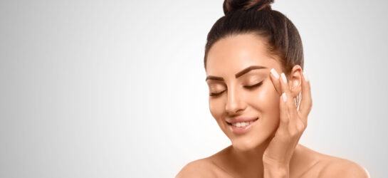 Junge Frau mit gepflegter Haut lächelt entspannt und berührt sanft ihre Augenpartie – Darstellung von Beauty- und Anti-Aging-Tipps rund um Augenfalten.