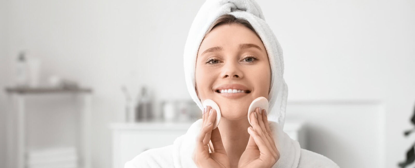 Auf dem Bild sieht man eine junge Frau in einem weißen Bademantel mit Handtuch-Turban, die sanft zwei Wattepads über ihre Wangen führt. Sie lächelt entspannt, während sie ihre Hautpflegeroutine durchführt. Das Bild zeigt ideal, wie richtiges Abschminken funktioniert: behutsam, gründlich und in einer ruhigen Atmosphäre. Der Hintergrund ist hell und minimalistisch – perfekt, um den Fokus auf die Reinigung des Gesichts zu legen.