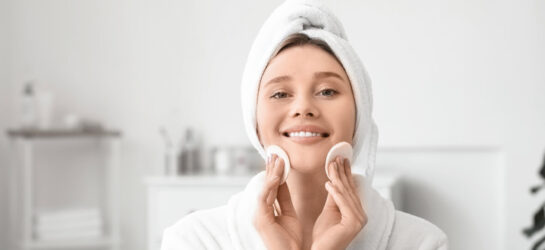Auf dem Bild sieht man eine junge Frau in einem weißen Bademantel mit Handtuch-Turban, die sanft zwei Wattepads über ihre Wangen führt. Sie lächelt entspannt, während sie ihre Hautpflegeroutine durchführt. Das Bild zeigt ideal, wie richtiges Abschminken funktioniert: behutsam, gründlich und in einer ruhigen Atmosphäre. Der Hintergrund ist hell und minimalistisch – perfekt, um den Fokus auf die Reinigung des Gesichts zu legen.