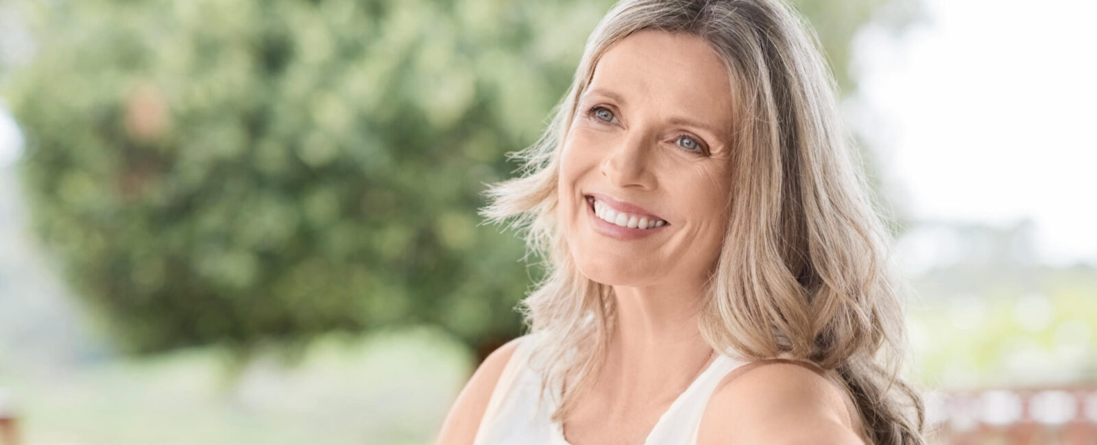 Auf diesem Bild sieht man eine Frau die ungeschminkt natürlich schön ist. Sie sitzt draußen in der Natur und lächelt.