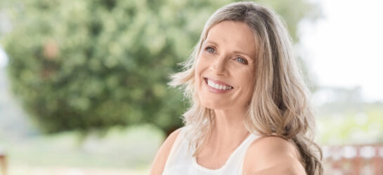 Auf diesem Bild sieht man eine Frau die ungeschminkt natürlich schön ist. Sie sitzt draußen in der Natur und lächelt.