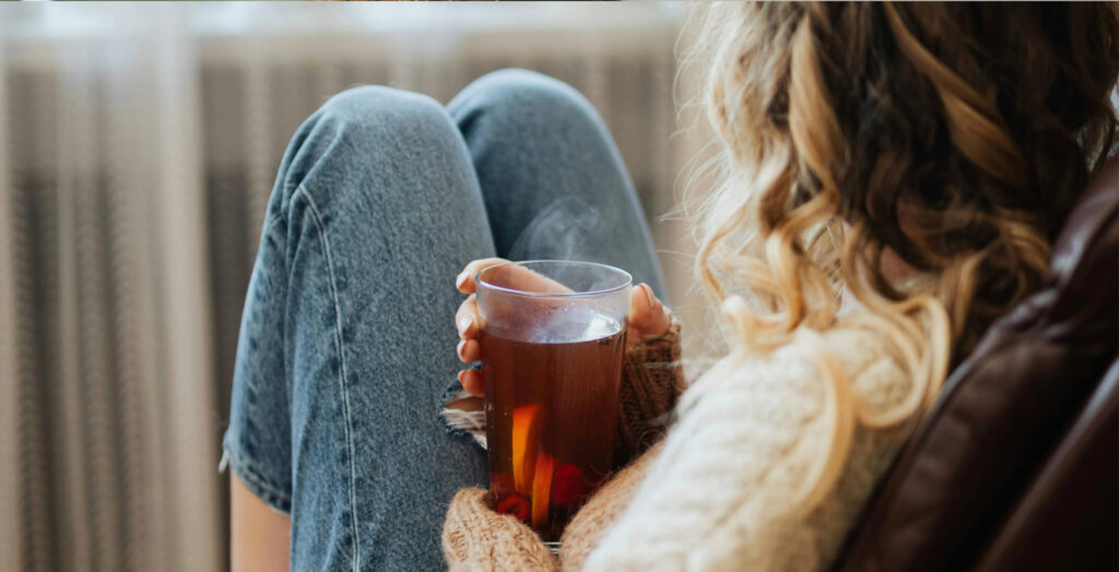 Auf dem Bild sitzt eine Frau mit einem warmen Getränk in der Hand. Das Bild steht somit für ein angenehmes Raumklima.